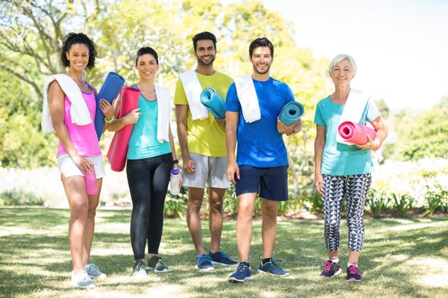 Group of diverse individuals standing in a park, each holding a rolled exercise mat and smiling. They are dressed in casual fitness attire, suggesting they are ready for an outdoor workout or yoga session. This image can be used for promoting fitness classes, outdoor activities, healthy lifestyle campaigns, or community events.