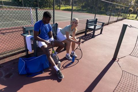 Tennis partners resting between sets on sunlit court holding rackets and water bottles