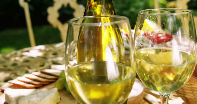 The image depicts an outdoor picnic setup with two glasses of white wine, a wine bottle, cheese, crackers, and fruits placed on a rustic table. The scene is sunny, creating a relaxed and inviting atmosphere. This image can be used for advertisements, social media posts, or websites focusing on outdoor activities, social gatherings, summer events, food and drink, or lifestyle blogs.