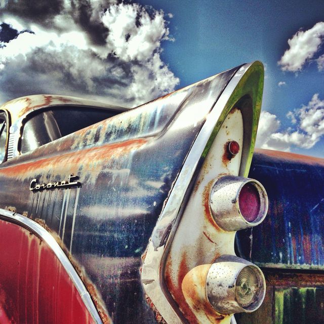 The image depicts the rear corner of a vintage car with weathered paint and rust, contrasted against a dramatic cloudy sky. The close-up view highlights chrome details and the unique design of the classic car. This image can be used in projects related to automotive restoration, nostalgia, history of automobiles, and vintage aesthetics.
