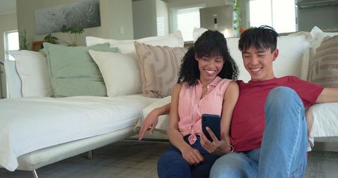 Multicultural couple relaxing on floor leaning against white sofa checking smartphone
