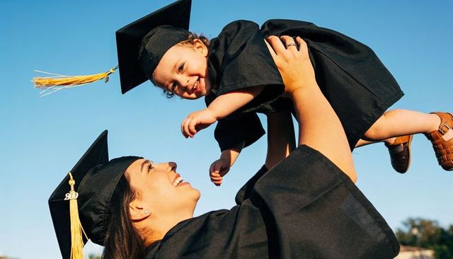 Graduate mother lifting toddler in cap and gown celebrating graduation outdoors on campus