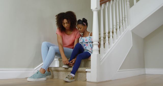 Happy african american mother with daughter sitting on stairs and tying shoes at home. Family, domestic life, education and lifestyle, unaltered.
