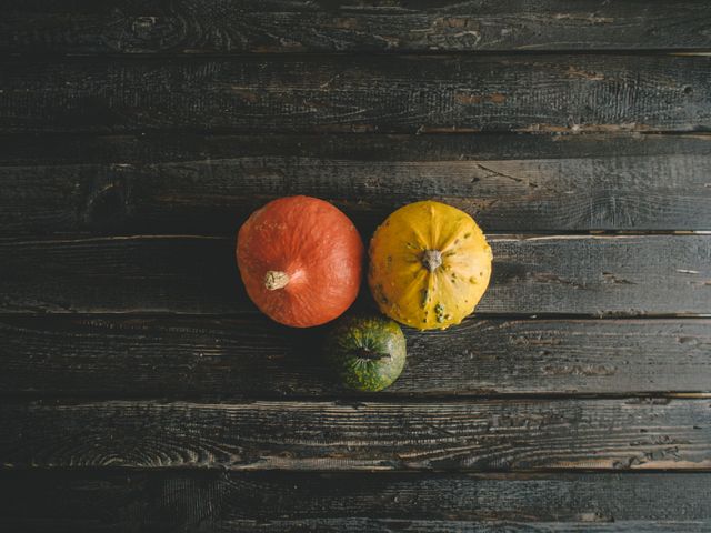 Three pumpkins of different colors are arranged on a dark, weathered wooden surface. The vibrant colors and rustic background create a striking contrast. This image can be used for autumn-themed projects, harvest festival promotions, seasonal decorations, or agricultural content.