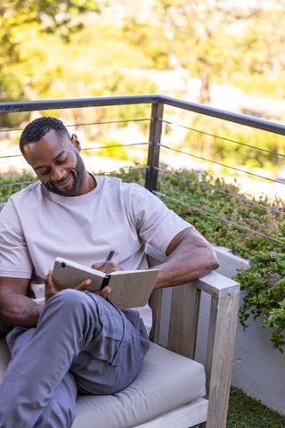 African man sitting on balcony journaling and writing in notebook in sunlight