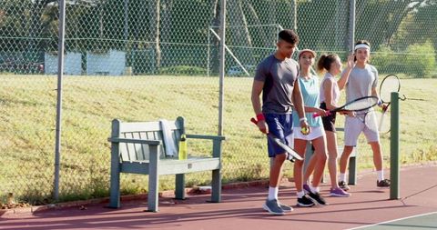 Sunlit diverse teens walking on outdoor tennis court holding rackets and balls, casual sports scene