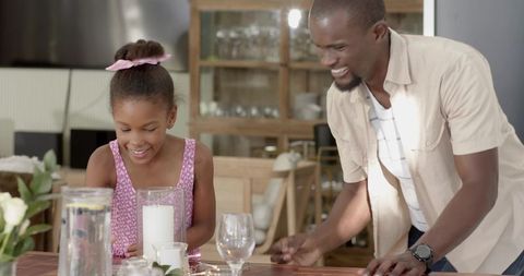 Smiling diverse father and daughter arranging cozy candlelit dining table for family