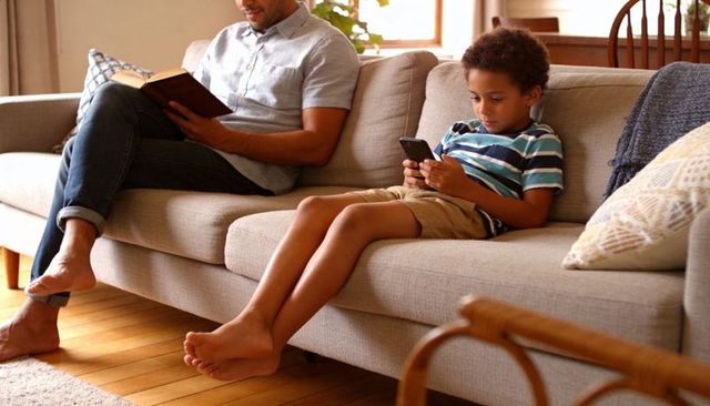 Father reading while son scrolling smartphone on sunlit couch during cozy family moment
