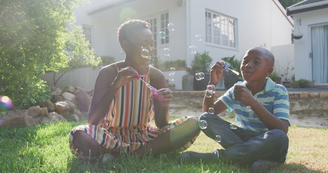 Happy african american mother and son sitting on grass and blowing bubbles in garden. Lifestyle, domestic life, family, and togetherness.