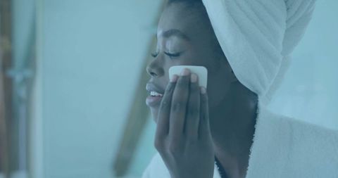 Woman cleansing face with cotton pad, towel turban and bathrobe, closeup skincare