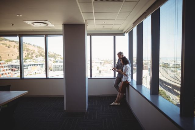 Executives standing near large office windows, engaging in a discussion while using a laptop. Sunlight streams in, illuminating the modern workspace. This image is ideal for promoting business collaboration, corporate meetings, teamwork, and professional environments.