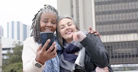 Smiling senior women taking selfie on city plaza in scarves and jackets, candid friendship