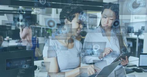 Two women collaborating on tablet and laptop with futuristic holographic interface overlays