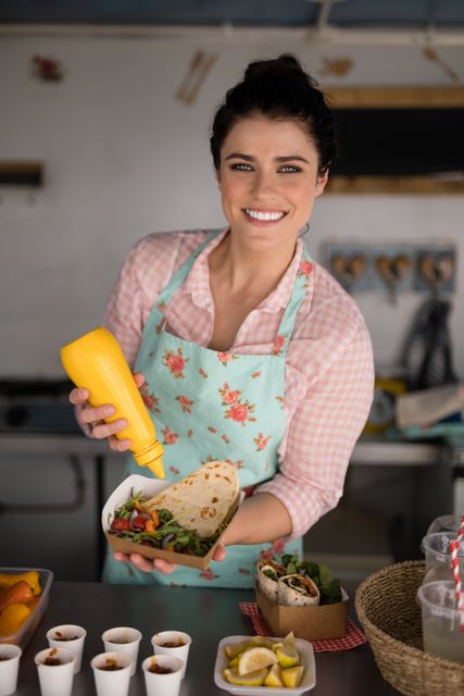 Waitress in a food truck smiling while preparing a meal, wearing a floral apron. Ideal for use in articles or advertisements related to street food, food trucks, customer service, and the hospitality industry. Can also be used for promoting casual dining experiences and fresh food preparation.