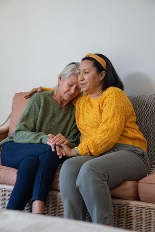 Comforting senior women holding hands on woven sofa showing compassionate friendship