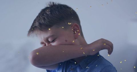 Young boy sneezing into elbow in soft pastel bedroom with gold star confetti