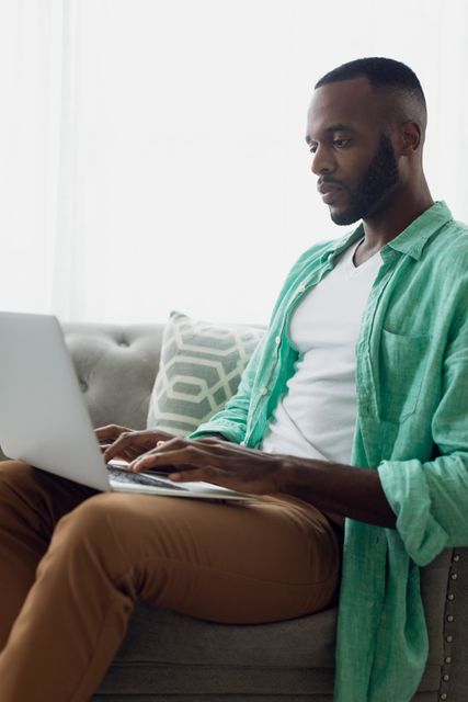 Side view of an African-American man sitting on a grey couch with white curtains inside a room while using a laptop
