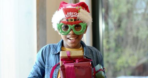 Preteen wearing santa hat and green novelty glasses holding stacked wrapped holiday gifts