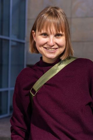 Smiling young woman standing at modern building wearing burgundy sweater and olive strap