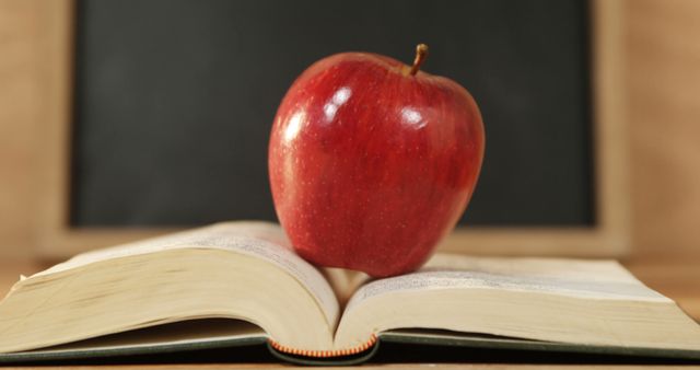 A red apple sits atop an open book in front of a chalkboard, symbolizing education and learning. It evokes the traditional connection between apples and teaching, often representing a gift to a teacher or a love for knowledge.