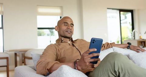 Mature man reclining on sofa checking blue smartphone in bright minimalist living room
