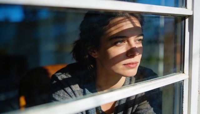 Woman gazing through sunlit window with striped shadows, thoughtful home interior portrait