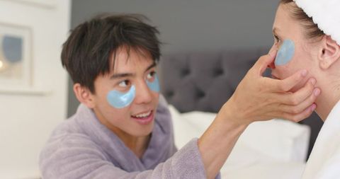 Couple applying blue hydrogel under-eye patches on bed robes sharing cozy skincare ritual