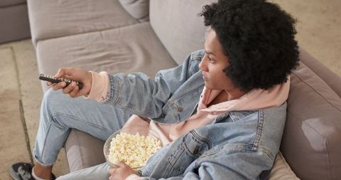 Young african american man relaxing on couch watching tv holding remote and popcorn