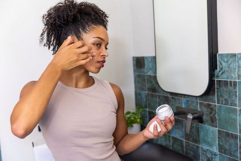 African american woman applying moisturizer at bathroom sink holding jar and checking skin