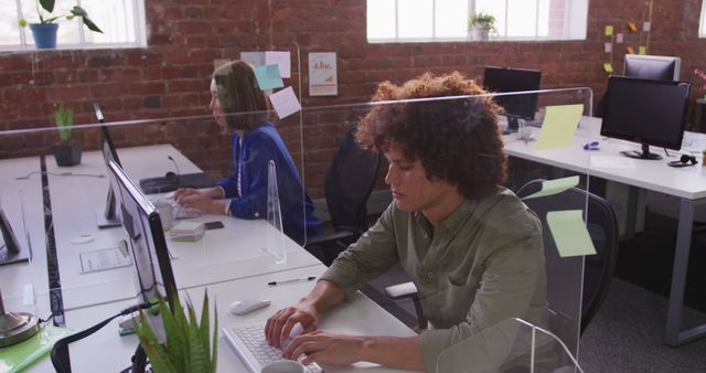 Diverse male and female business colleagues sitting at desks with sneeze guard using computers. business in a modern office.