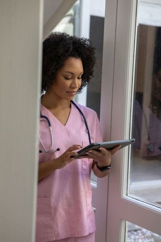 African American Nurse in Pink Scrubs Using Tablet by Glass Door