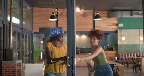 Two women entering trendy cafe pushing glass doors under open sign sharing laughter