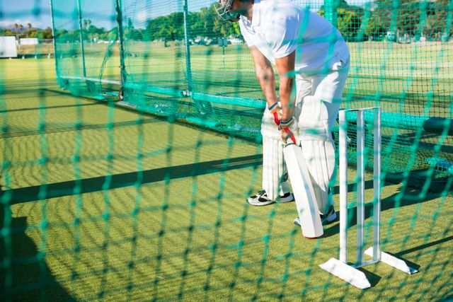 Cricketer practicing batting in nets on a sunny day. Ideal for use in sports-related content, training guides, fitness blogs, and recreational activity promotions.