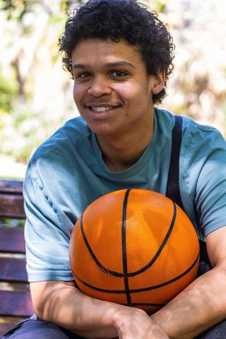 Smiling african american young man holding basketball on sunlit park bench
