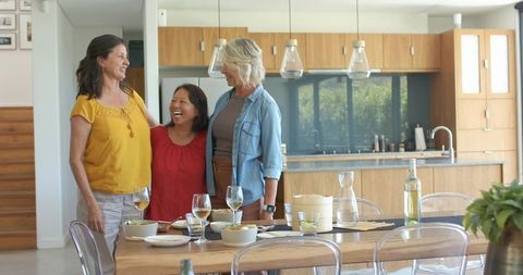 Multicultural women laughing around wooden dining table in modern open-plan kitchen with wine