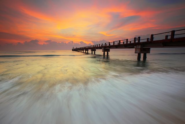 Image depicts a tranquil scene of a pier extending over the ocean while the sky is illuminated with vibrant sunset colors. The gentle waves create a serene atmosphere. This image can be used for travel promotions, relaxation and wellness concepts, beach resort advertising, or any project looking to evoke a sense of calm and beauty.