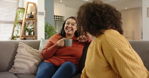 Two friends laughing and chatting on cozy gray sofa over coffee in bright living room