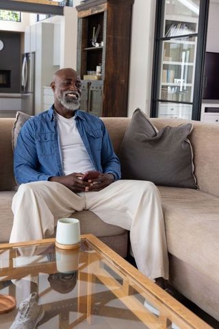 Senior african american man relaxing on sofa holding smartphone in modern sunlit living room