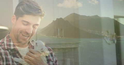 Smiling man cradling small white dog with coastal reflection through sunlit window