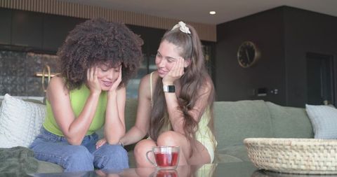 Diverse female friends sharing intimate conversation on green sofa in modern living room