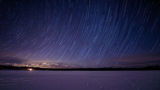 Arcing star trails over frozen alpine lake with distant cabin lights
