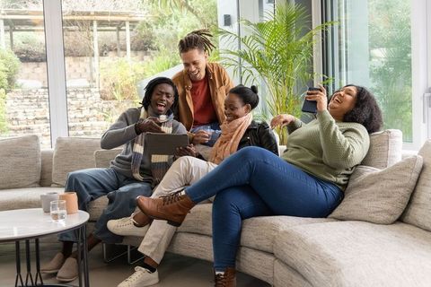 Diverse friends laughing and watching tablet while relaxing on light-gray sectional sofa