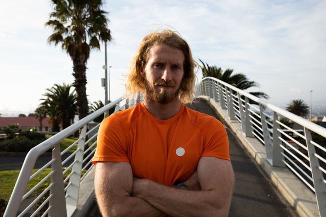 Portrait of a fit Caucasian man with long blonde hair wearing sportswear exercising outdoors in the city on a sunny day with blue sky, looking straight to the camera standing on footbridge with arms crossed.
