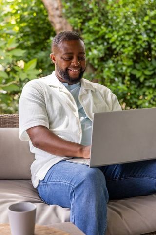 African man smiling and working remotely on laptop on patio sofa in lush garden