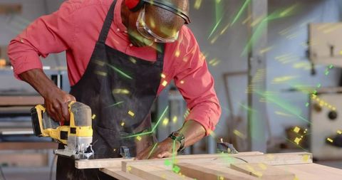 Woodworker cutting plank with handheld jigsaw in workshop wearing apron and safety goggles