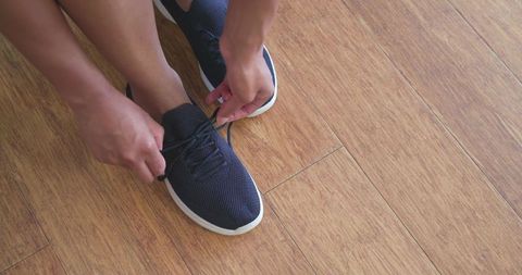 African american woman tying navy sneaker laces on light hardwood floor closeup detail