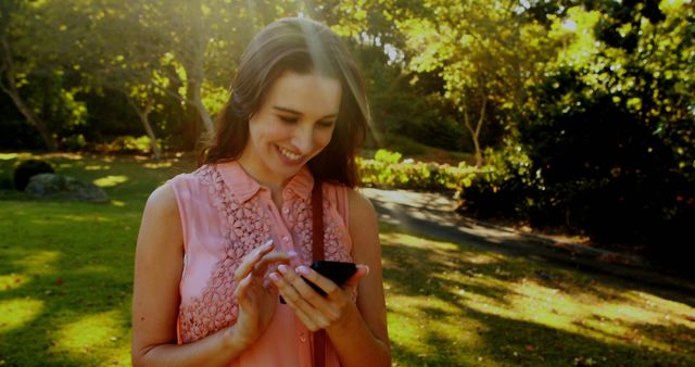 Woman engaging with smartphone while enjoying sunny day in park. Suitable for themes of technology, leisure, outdoor activities, and social connection. Perfect for advertising apps, lifestyle websites, and promotional materials focusing on connectivity and youth culture.