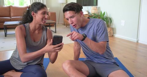 African american friends sharing workout app on phone while sitting on yoga mats at home