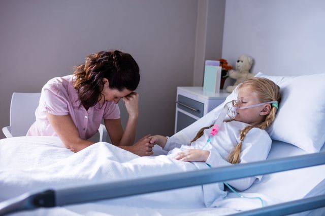 Mother holding hand of her hospitalized child, who is lying in a hospital bed with an oxygen mask. The mother appears worried and emotional, providing support to her sick child. This image can be used in contexts related to healthcare, family support, pediatric care, and emotional moments in medical settings.