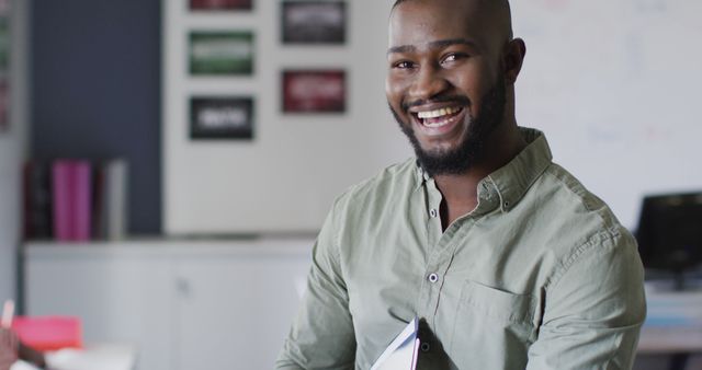 African American man in a green shirt is confidently smiling in an office. He holds a notebook, suggesting he is ready for a meeting or presentation. The background includes office furniture and decorations. Useful for themes related to business, professionalism, workplace dynamics, and positive work environment.